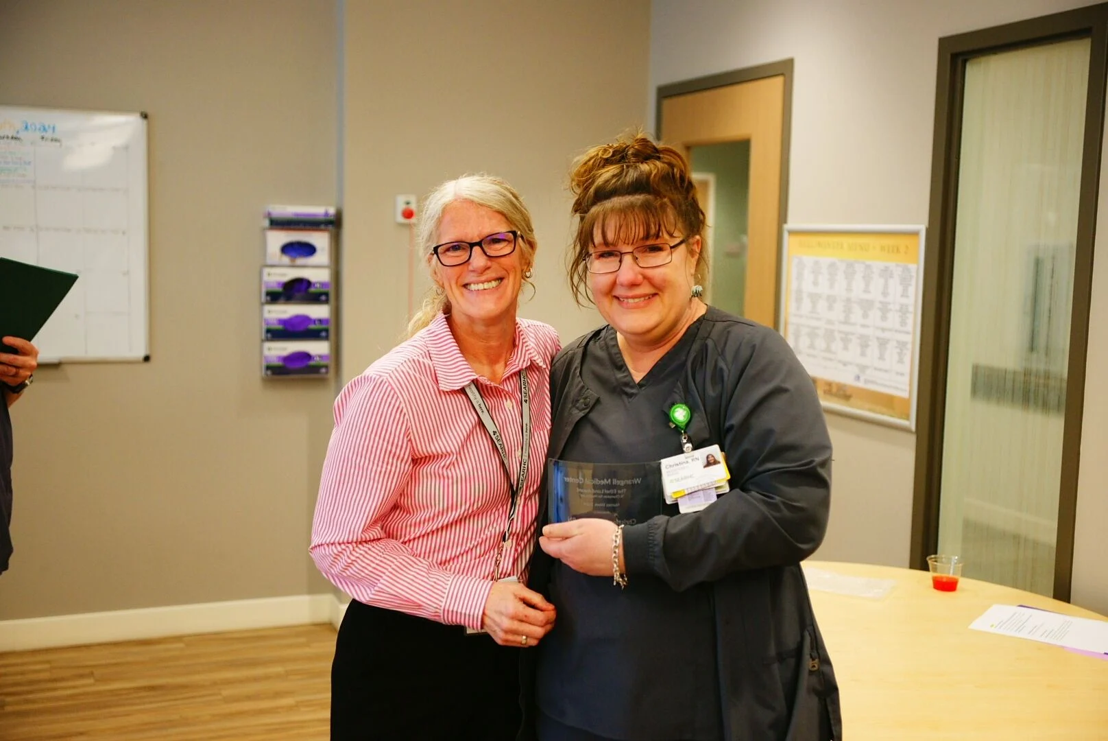 Two smiling women, one in a striped shirt and one in scrubs, holding a clear award plaque in an office setting.