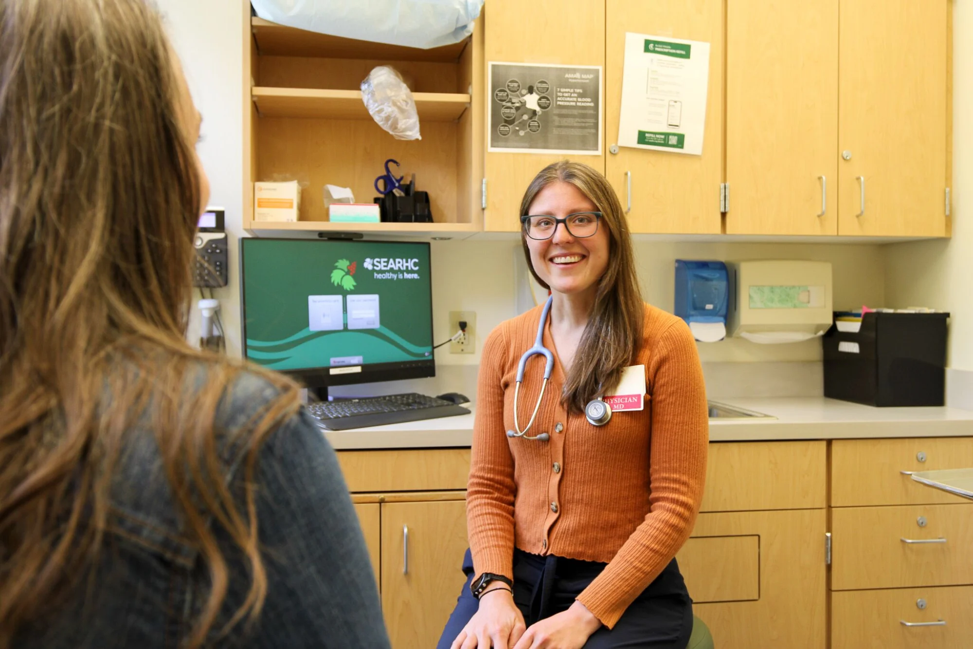A smiling female physician with a stethoscope and name tag looks at a patient in a medical exam room.