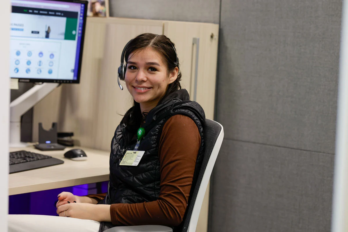 A young woman wearing a headset and ID badge smiles at the camera while seated at a computer desk in an office.