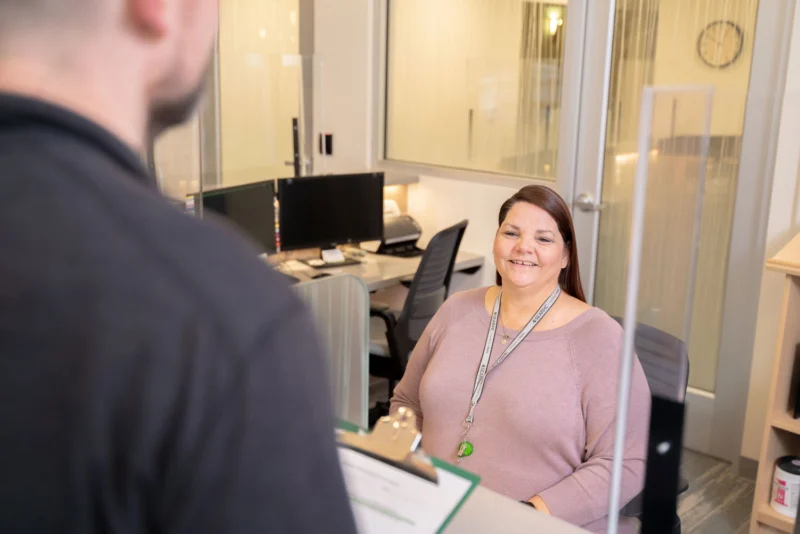 A smiling woman in a pink shirt sits at an office desk, looking at a person holding a clipboard in the foreground.
