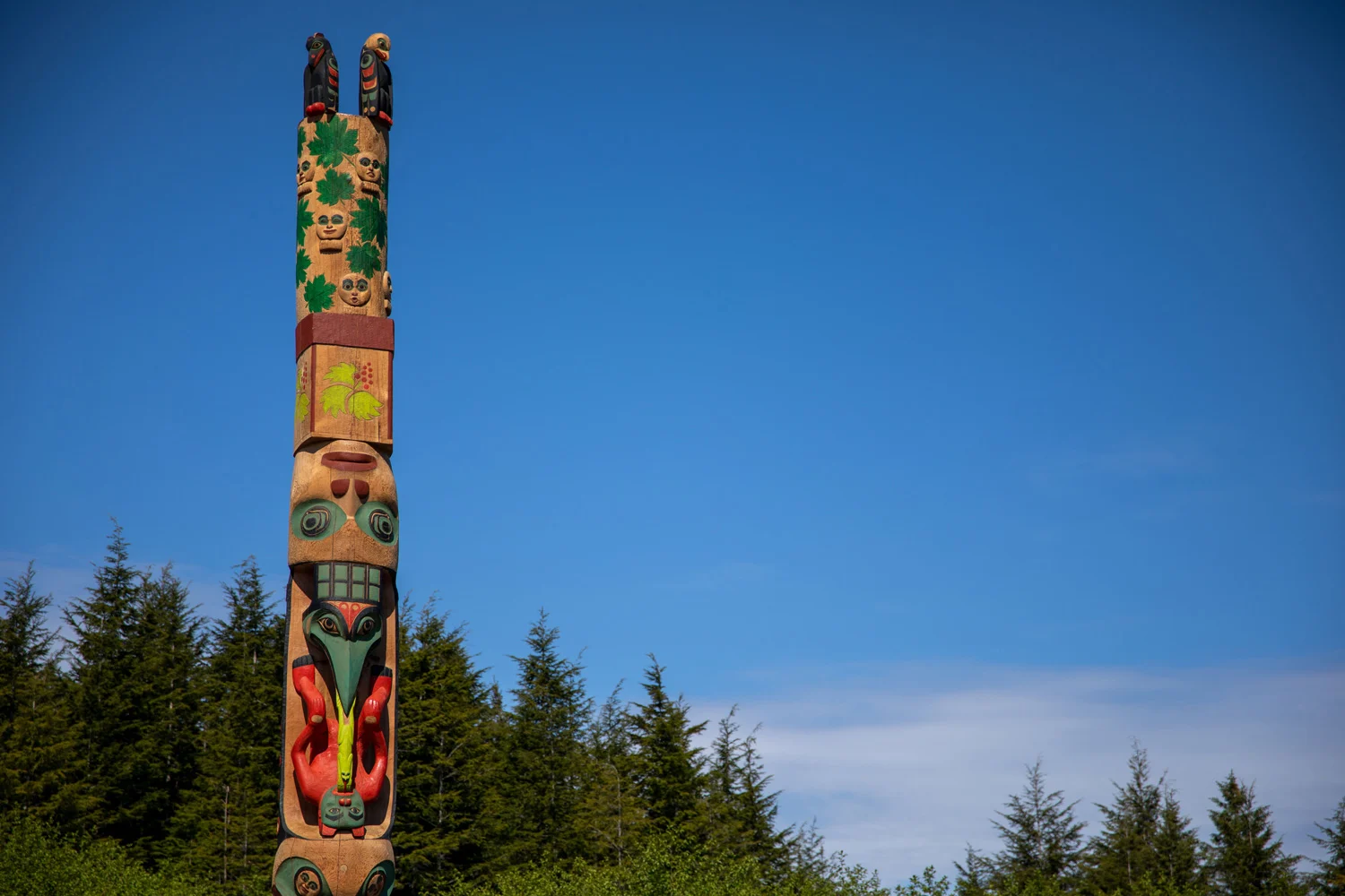 A tall, colorful totem pole with multiple carved figures and designs, including birds, faces, and animals, set against a bright blue sky and a backdrop of dark green evergreen trees.