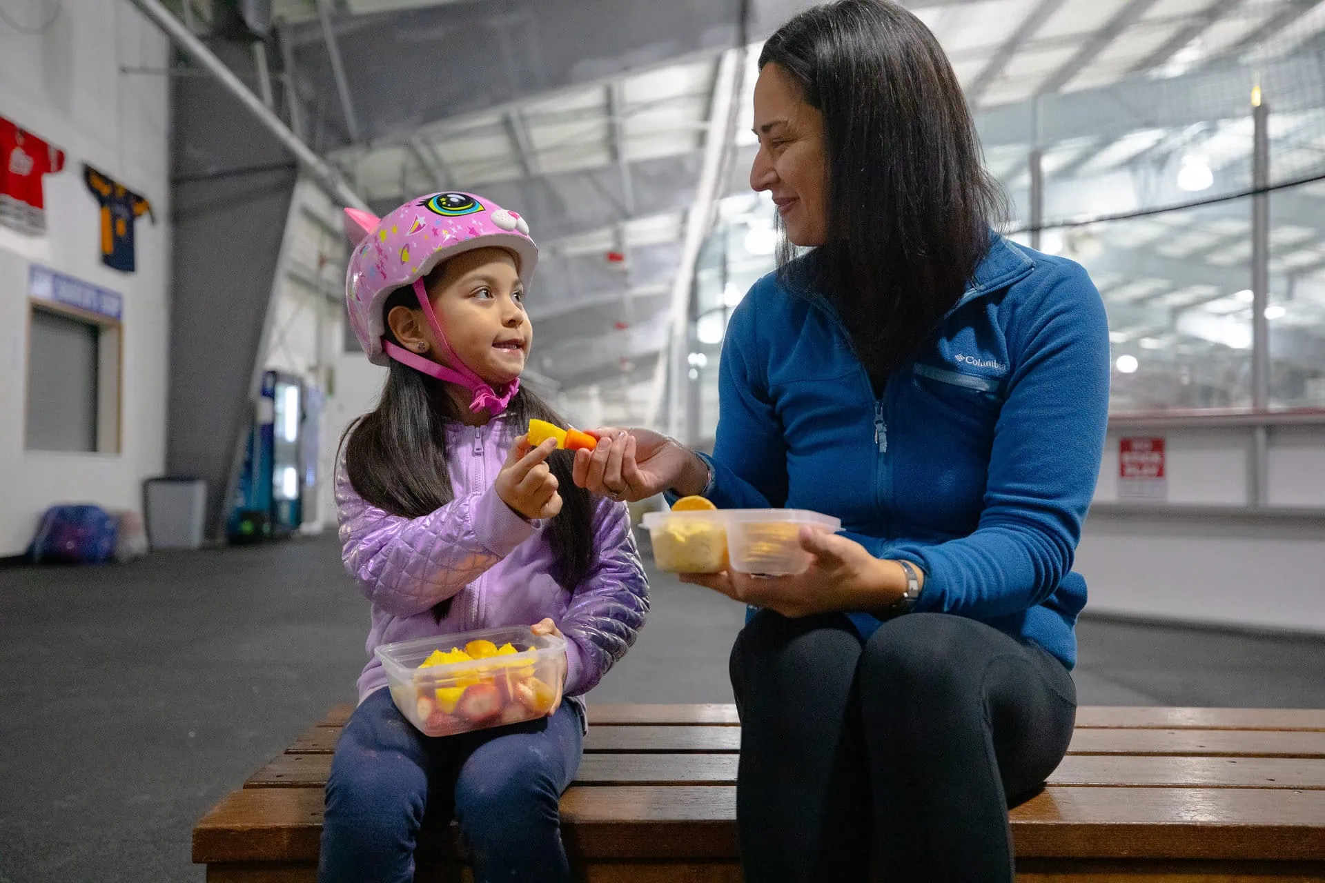 A woman and a young girl in a pink helmet share fruit and snacks on a bench in an indoor facility.