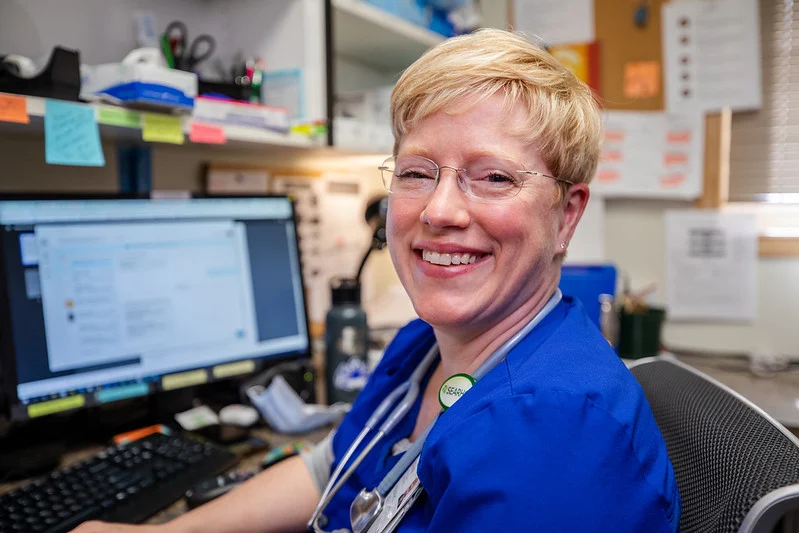 A smiling woman in blue scrubs and glasses sits at a desk with a computer and stethoscope.