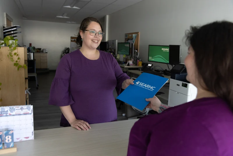 A woman in a purple shirt hands a blue folder with the SEARHC logo to another person across a counter in an office setting.