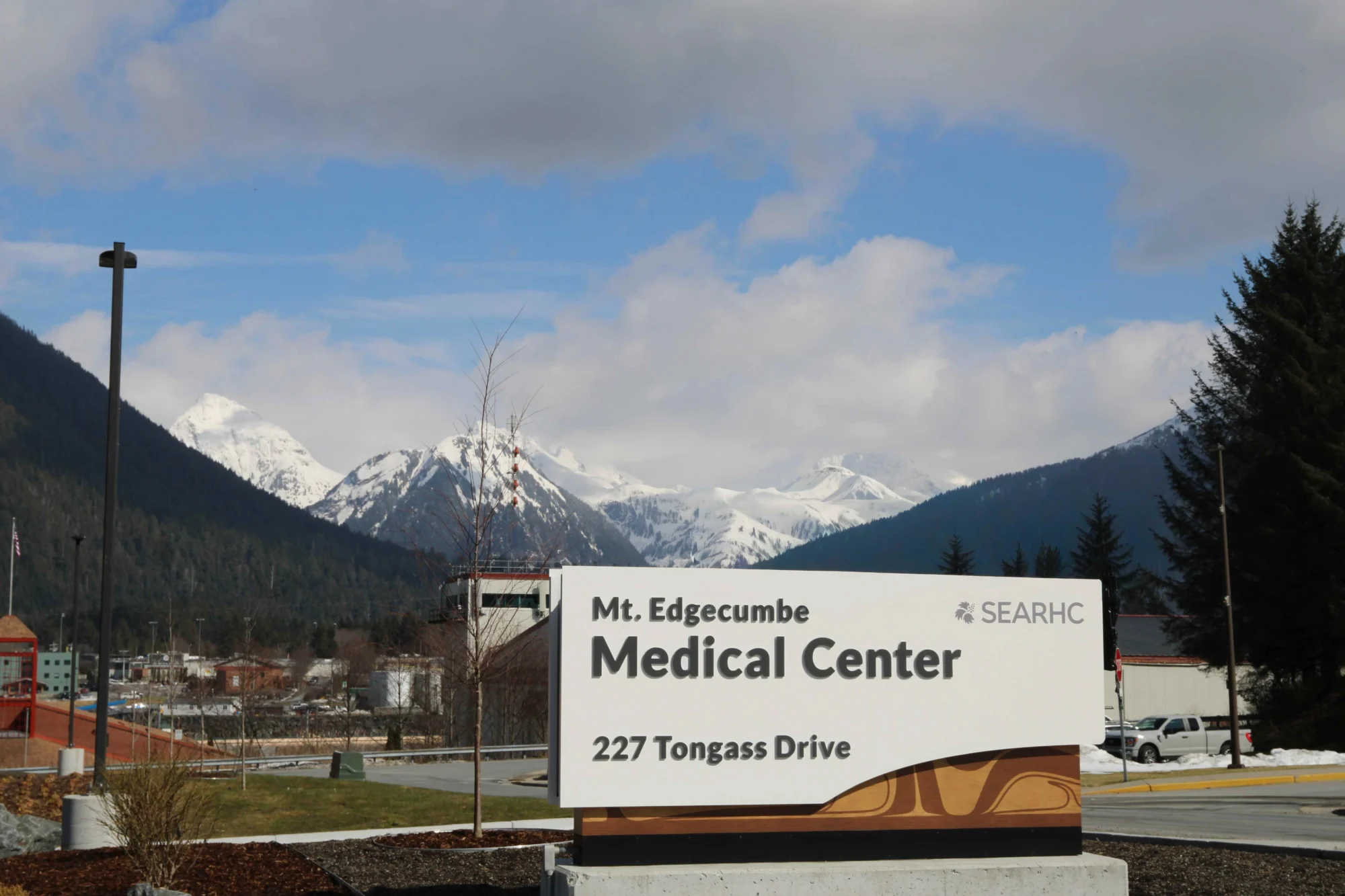 A white sign for Mt. Edgecumbe Medical Center with its address, set against a backdrop of snow-capped mountains, forested hills, and a blue sky with clouds.