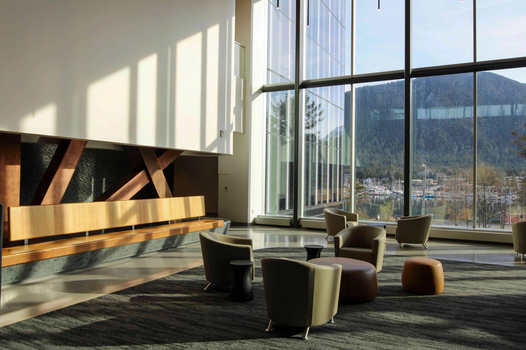 A modern lounge with large windows overlooking a marina, water, and forested mountains. The interior features a wooden bench, armchairs, and ottomans on a patterned rug, with sunlight casting shadows.