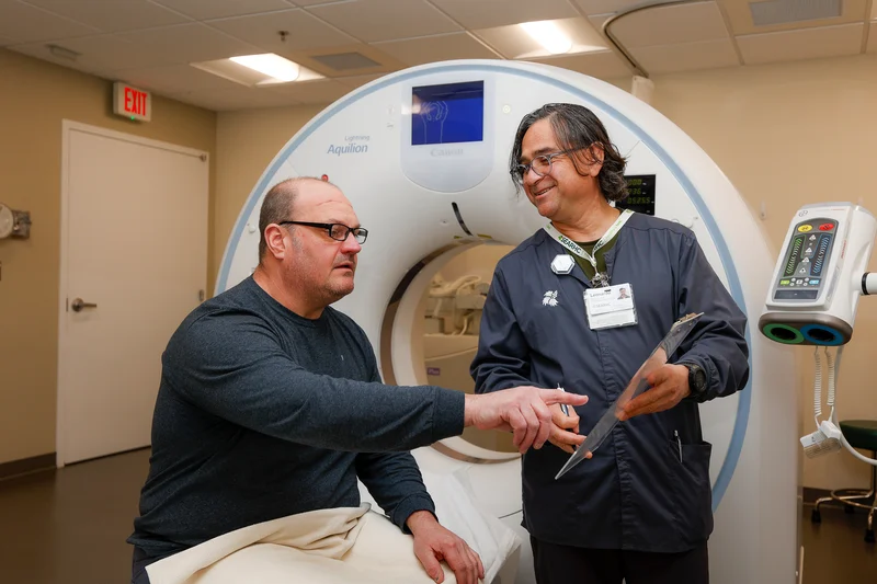 A patient and a medical professional discuss a procedure in a room with a large CT scanner.