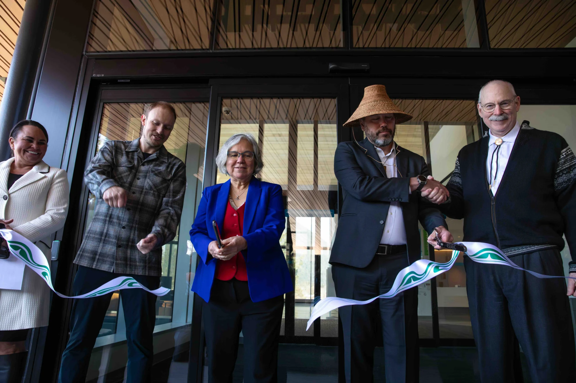 Five people, including two men holding scissors, cut a white ribbon with green designs in front of a modern glass building entrance.