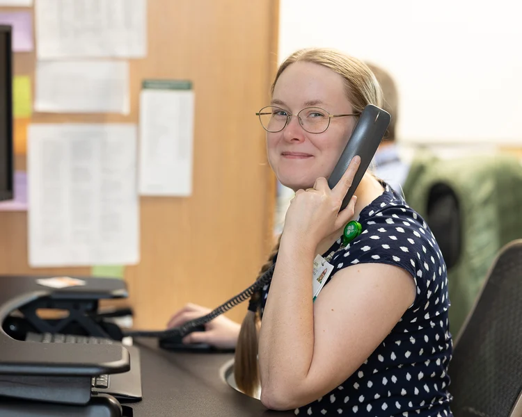 A smiling woman with glasses and a braided ponytail talks on a phone at a desk.