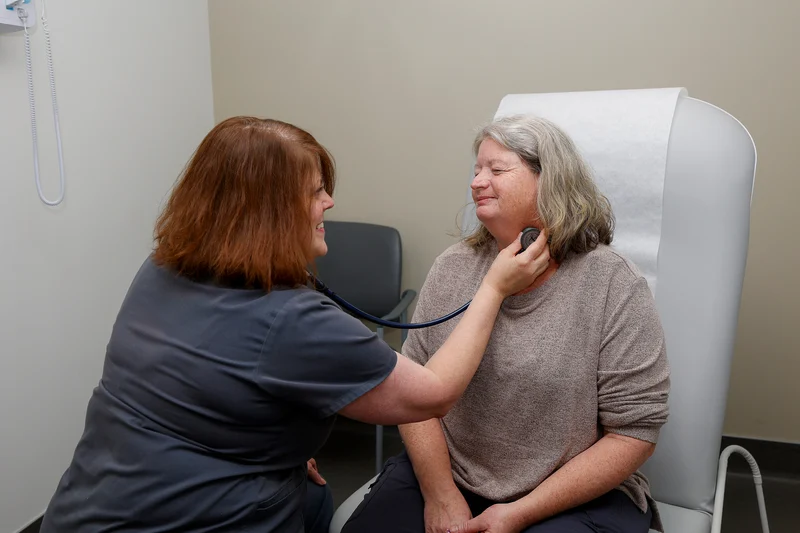 A medical professional uses a stethoscope on a smiling patient's neck during an examination.