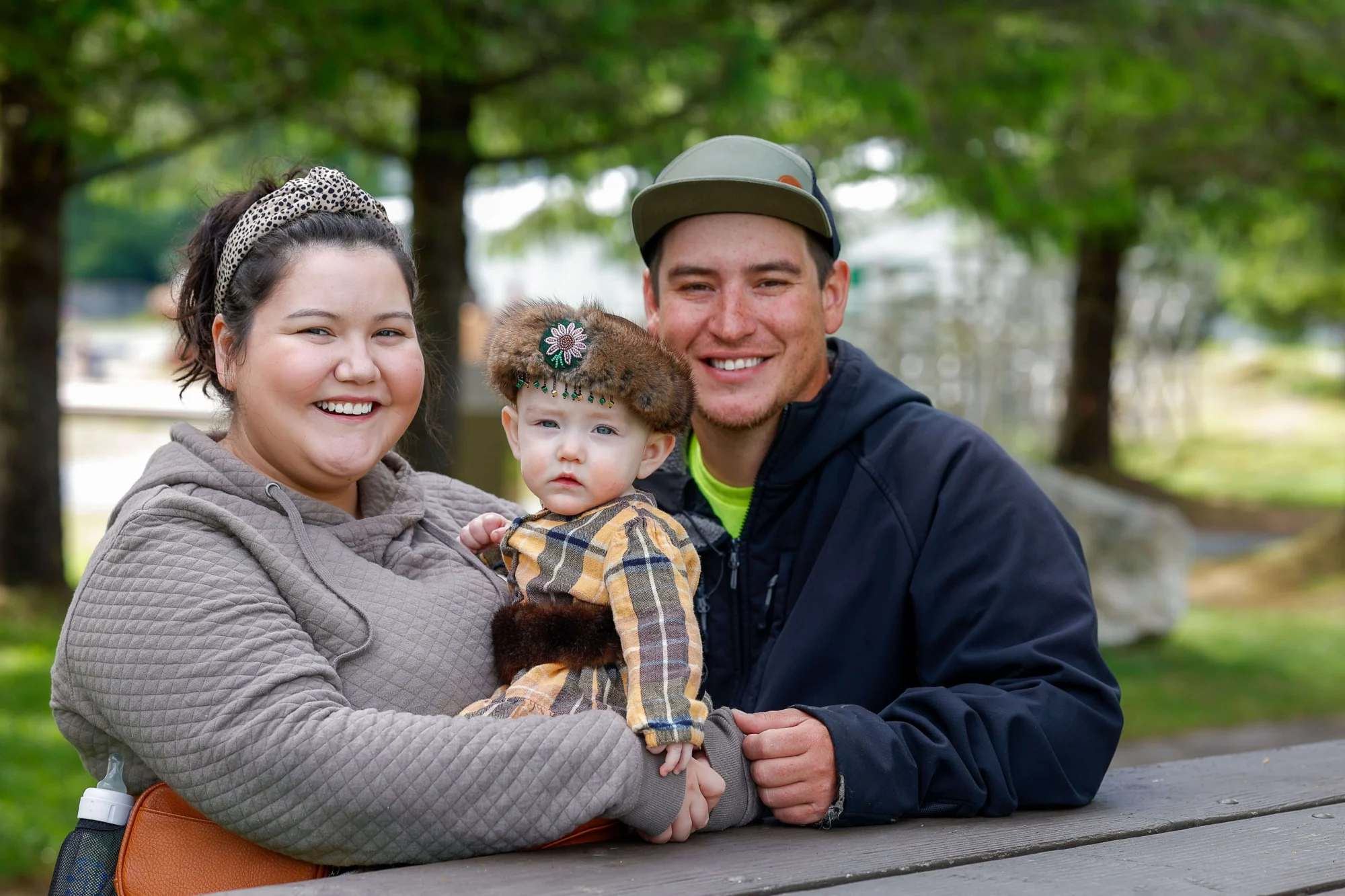 A family of three, a mother, father, and baby, smile at the camera while sitting at a picnic table outdoors. The baby wears a fur hat with beaded floral design.