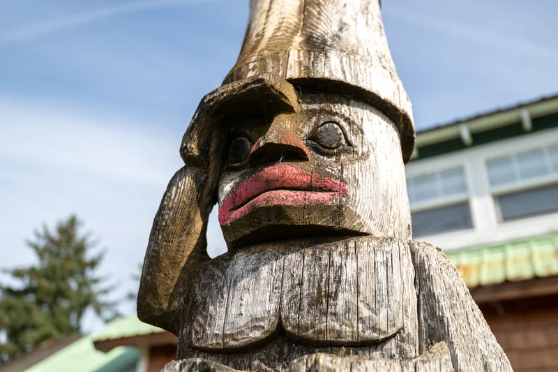 Close-up of a weathered wooden totem pole figure with a pink-painted mouth and a hand raised to its forehead, against a blurred sky and building.