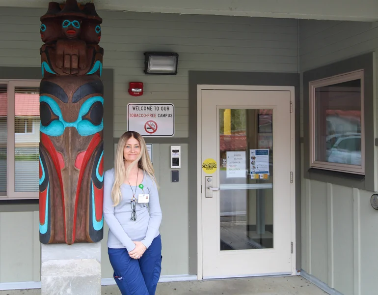 A woman in scrubs stands next to a colorful carved totem pole outside a building entrance with a 'Tobacco-Free Campus' sign.