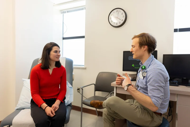 A male doctor in a checkered shirt and a female patient in a red top are talking in a bright medical examination room.