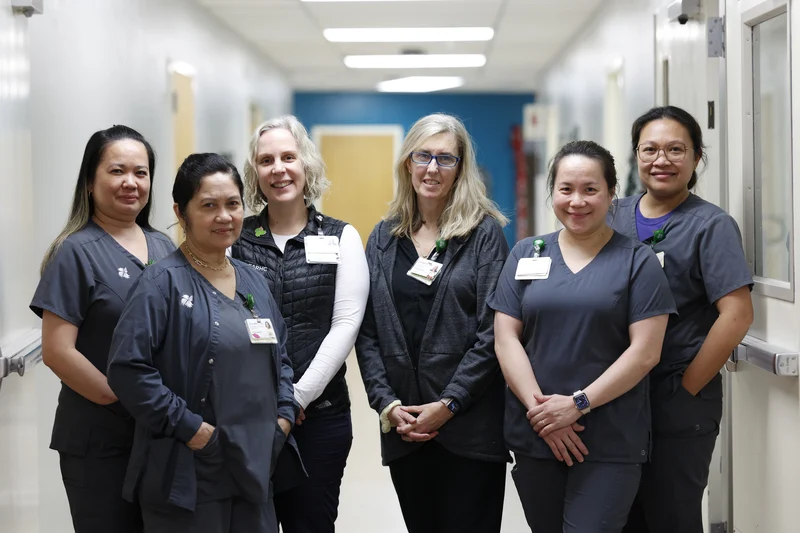 Six smiling women, mostly in grey scrubs, stand in a hospital hallway.