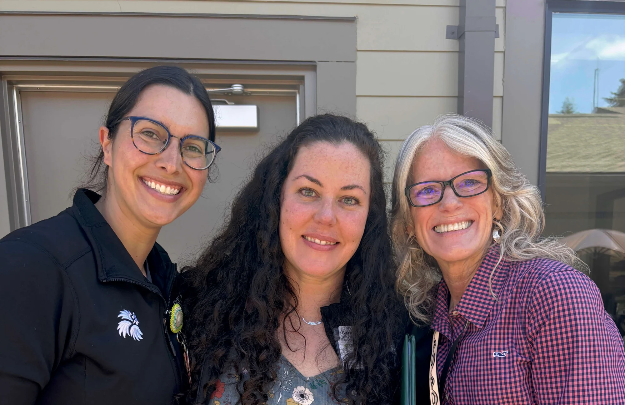 Three smiling women, one with dark hair and glasses, one with long curly hair, and one with gray hair and glasses, pose in front of a building.