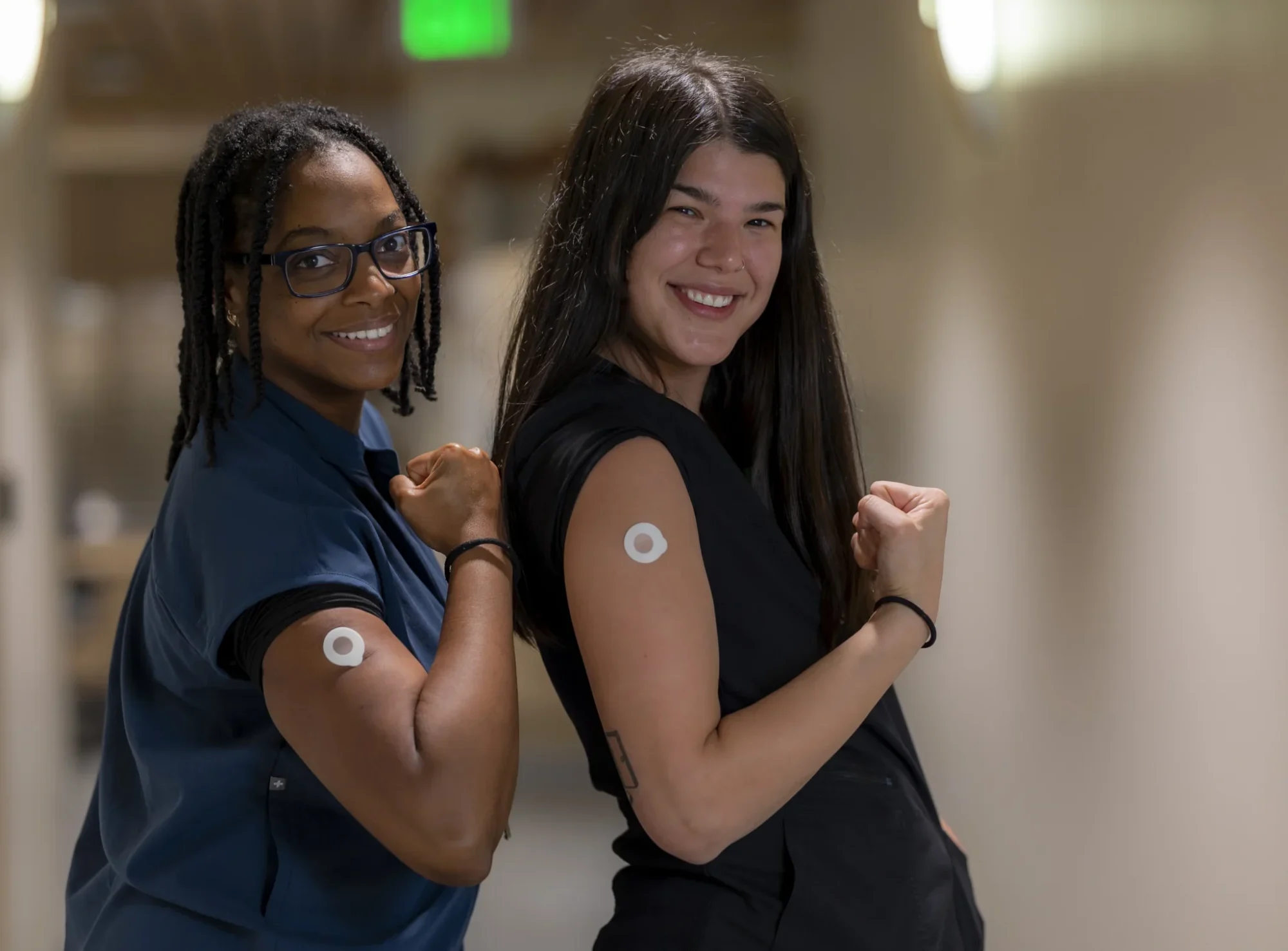 Two smiling women, one Black and one white, stand side-by-side in a hallway, flexing their biceps. Both have white circular continuous glucose monitors on their upper arms.